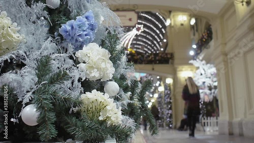 Christmas tree, decorated with decorative flowers, stands in the shopping center, against the background of passing visitors