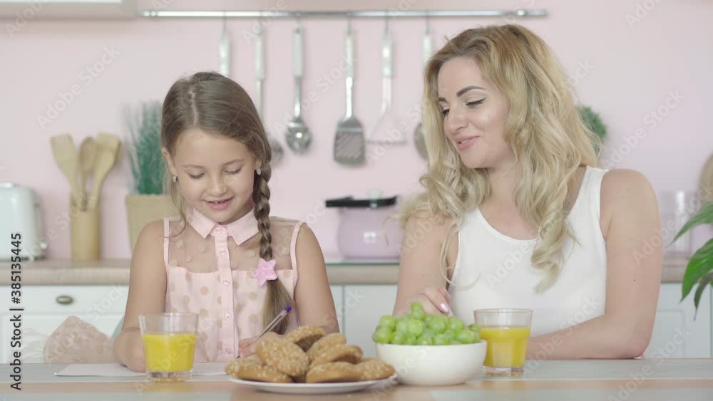 Beautiful young mother helping daughter with homework in the morning. Portrait of happy Caucasian parent and child sitting at the table in kitchen and talking. Beautiful young mother helping daughter with homework in the morning. Portrait of happy Caucasian parent and child sitting at the table in kitchen and talking.