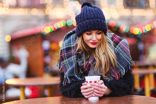Outdoors lifestyle fashion portrait of stunning blonde young woman. Drinking hot mulled wine and enjoying life. Wearing stylish fur coat, knitted hat and wide scarf. Romantic mood. Festive background 