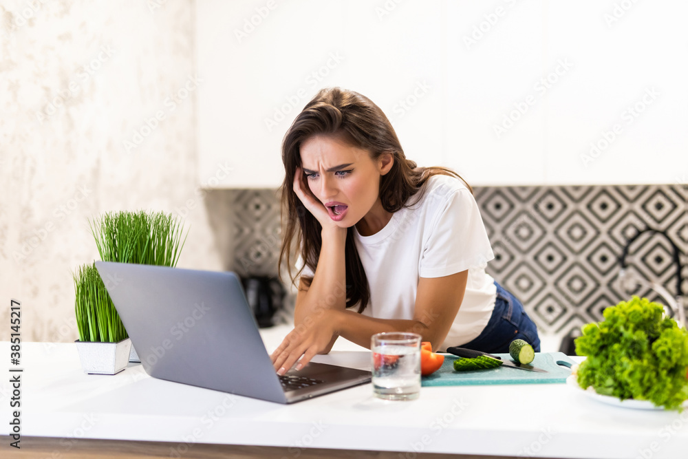 Happy young woman enjoying cooking looking for a recipe on the laptop in the kitchen Laptop on kitchen table and cooking girl. Food blogger concept Happy young woman enjoying cooking looking for a recipe on the laptop in the kitchen Laptop on kitchen table and cooking girl. Food blogger concept