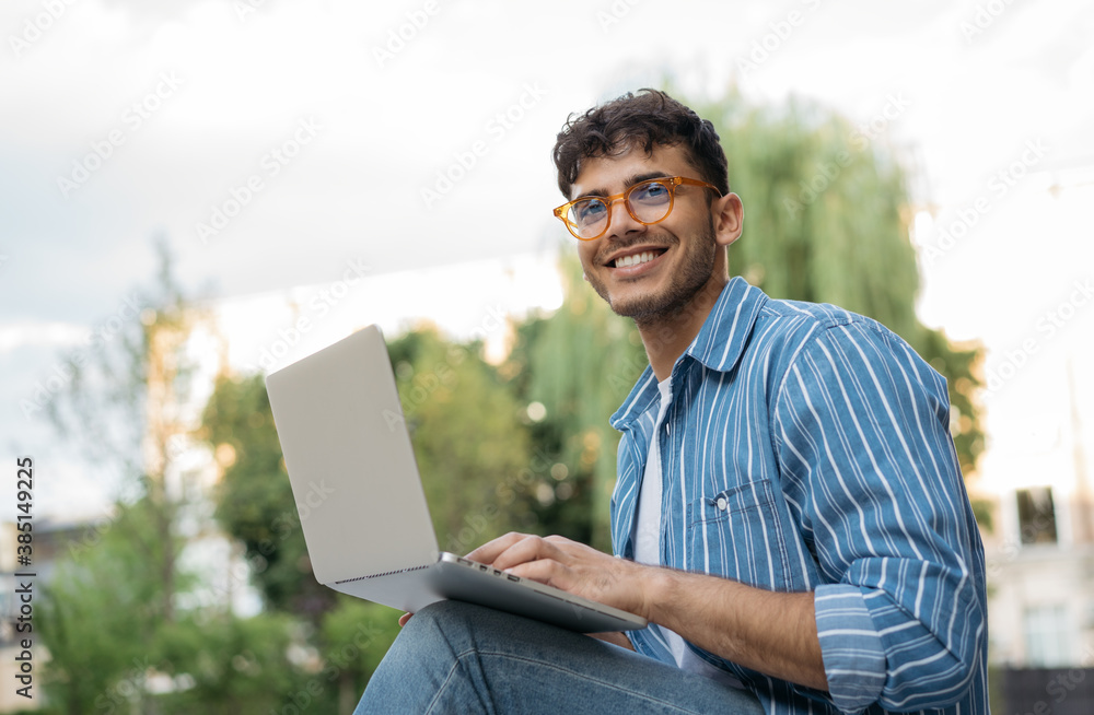 Portrait of handsome Indian copywriter using laptop computer, typing, working freelance project online, sitting outdoors. Smiling asian student studying, learning language, online education concept - obrazy, fototapety, plakaty 