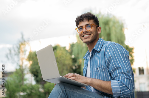 Portrait of handsome Indian copywriter using laptop computer, typing, working freelance project online, sitting outdoors. Smiling asian student studying, learning language, online education concept