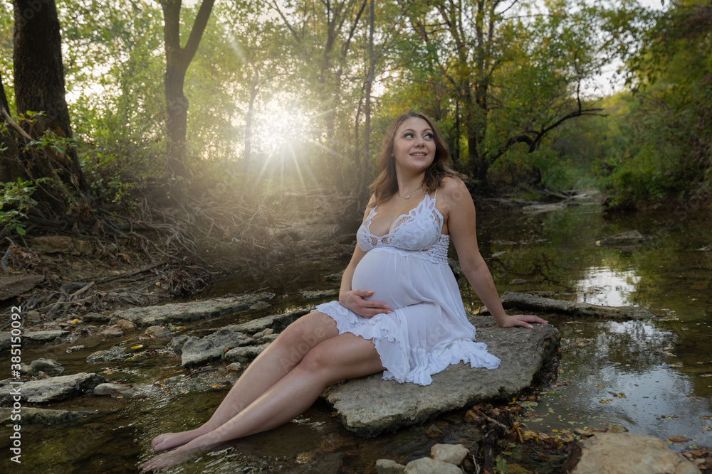 Fototapeta premium Young pregnant lady sitting on rock in forest creek hand on maternity belly beautiful outdoor portrait 