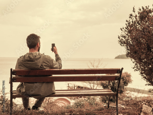 Man with back looking at phone screen while sitting on a bench outside