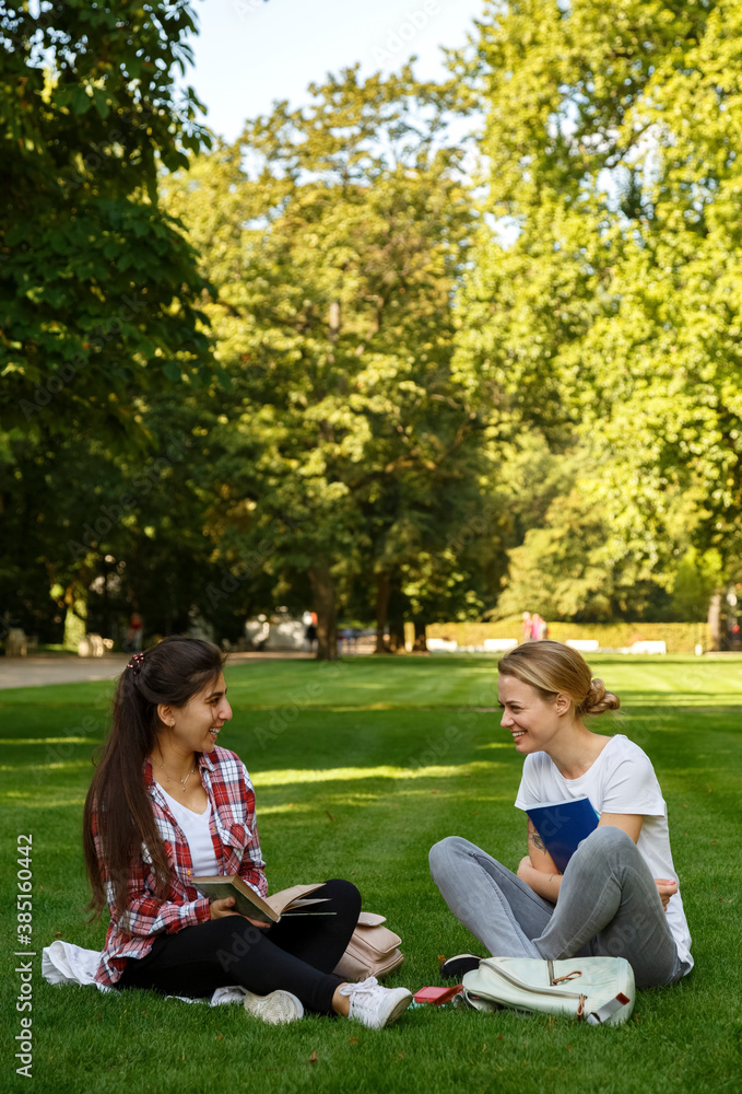 Laughing students with textbooks sitting on grass in green park Laughing students with textbooks sitting on grass in green park