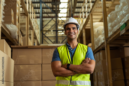 Portrait of young Indian industrial worker with arm folded working in logistic industry indoor inside factory warehouse. Smiling happy man in hard hat looking at camera arms crossed at depot