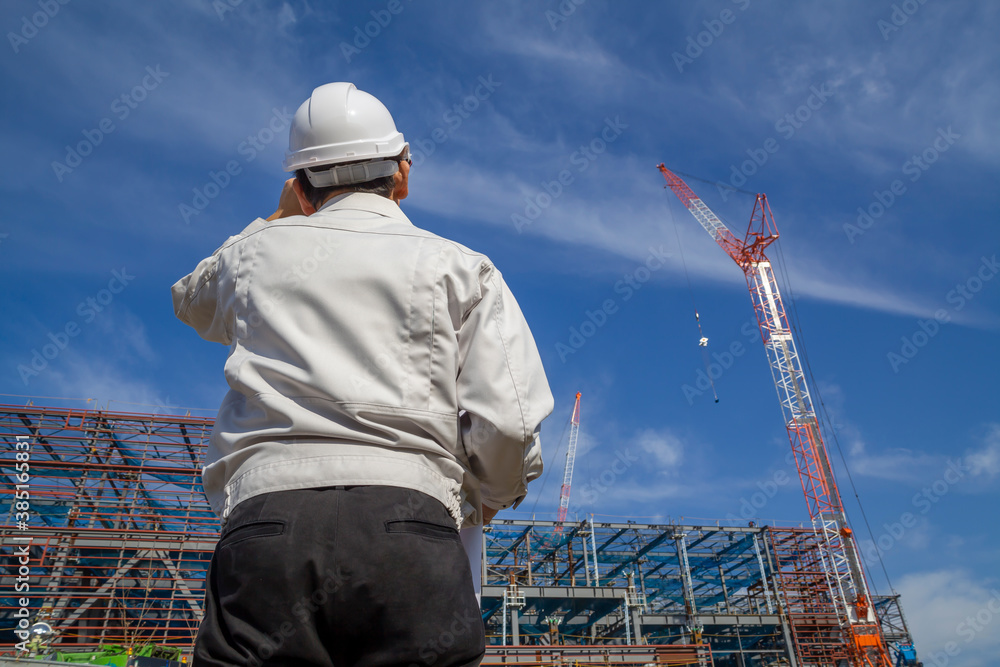 青空を背景にビル工事現場を見つめる作業着を着た男性技師の後ろ姿。