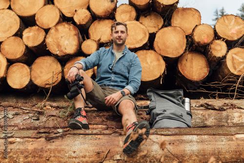 Man with binoculars and backpack sitting on log against woodpile in forest photo