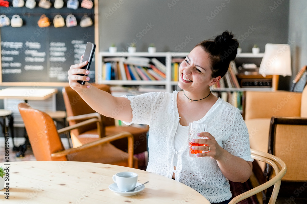 Smiling woman holding drink taking selfie with smart phone at table in cafe Smiling woman holding drink taking selfie with smart phone at table in cafe