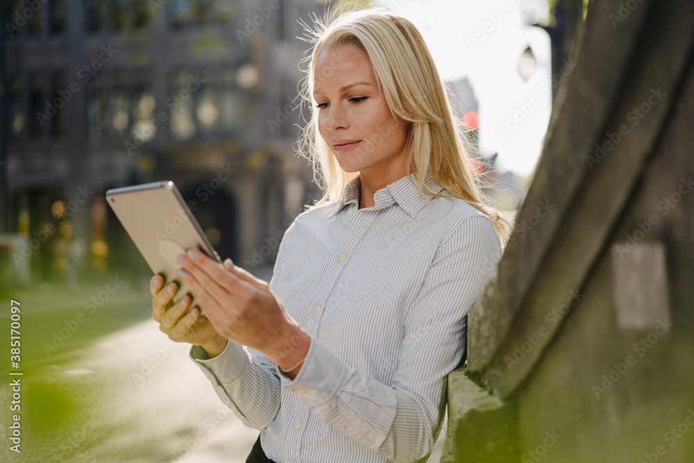 Blond beautiful female entrepreneur using digital tablet while leaning on wall in city Blond beautiful female entrepreneur using digital tablet while leaning on wall in city
