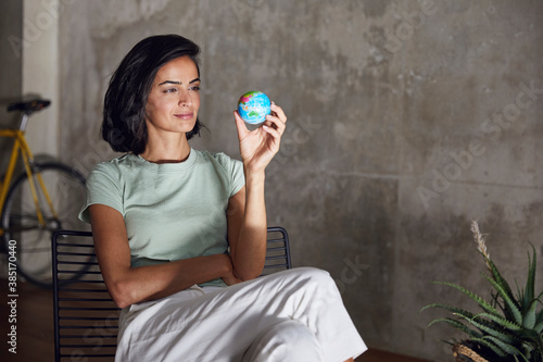 Businesswoman holding small globe while sitting on chair against wall in office photo