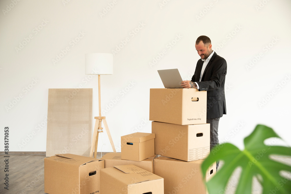 Businessman using laptop on cardboard box while standing in new unfurnished house Businessman using laptop on cardboard box while standing in new unfurnished house