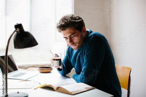 Male architect holding coffee reading book while sitting at desk in office