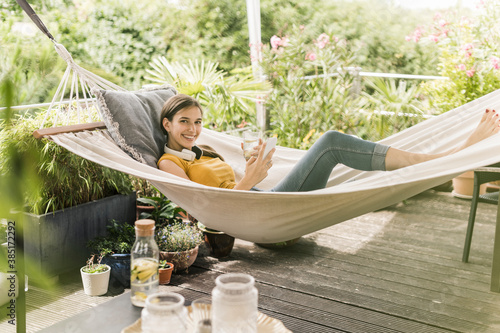 Smiling woman holding drink and smart phone while lying on hammock in yard photo