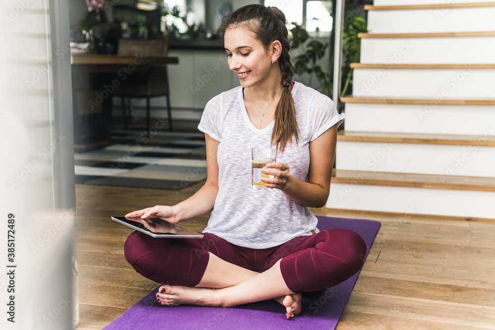 Smiling woman holding glass while using digital tablet on exercise mat at home Smiling woman holding glass while using digital tablet on exercise mat at home
