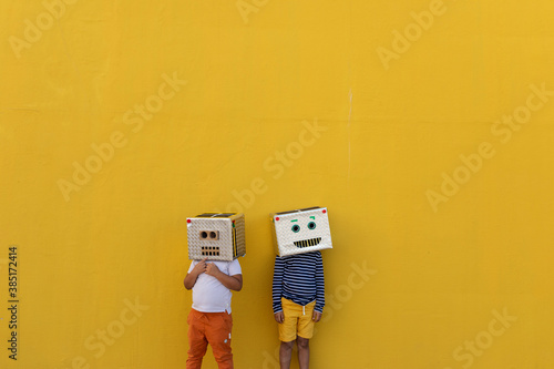 Boys wearing robot masks made of boxes while standing against yellow wall photo