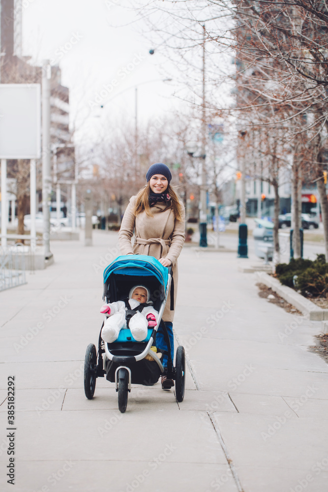 Fototapeta premium Happy young Caucasian mother walking with baby daughter outdoor in city. Family two people strolling together outside on city streets. Urban life with kids children.