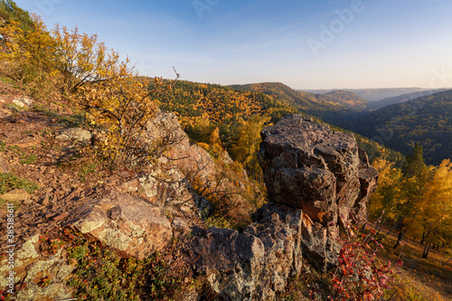 Autumn landscape in golden colors. In the foreground there are rocks, trees with yellow leaves. In the background, mountains covered with forest in the autumn haze. © Marina