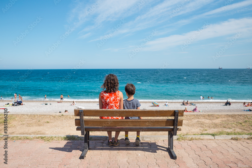 A woman with a child is sitting on a bench on the embankment and looking at the sea