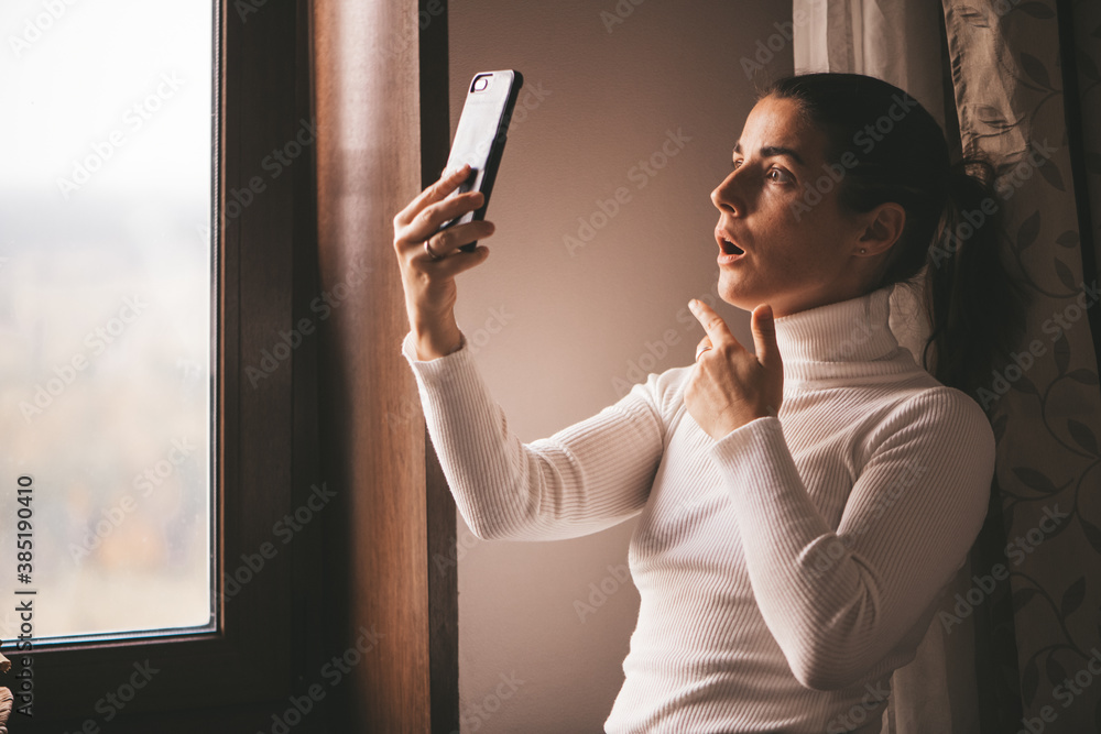 A shocked young woman in a white turtleneck looks at the screen of her phone at home in surprise. A stylish woman who has a video call on her smartphone. Video chat in social networks. A shocked young woman in a white turtleneck looks at the screen of her phone at home in surprise. A stylish woman who has a video call on her smartphone. Video chat in social networks.