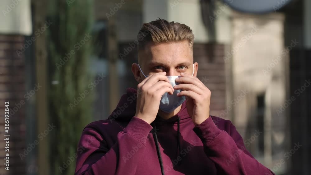 Portrait of a young man looking at the camera and putting on a medical mask to protect his face from the nCov-19 coronavirus. COVID-19 prevention measures. Slow motion. Face close up . Portrait of a young man looking at the camera and putting on a medical mask to protect his face from the nCov-19 coronavirus. COVID-19 prevention measures. Slow motion. Face close up .