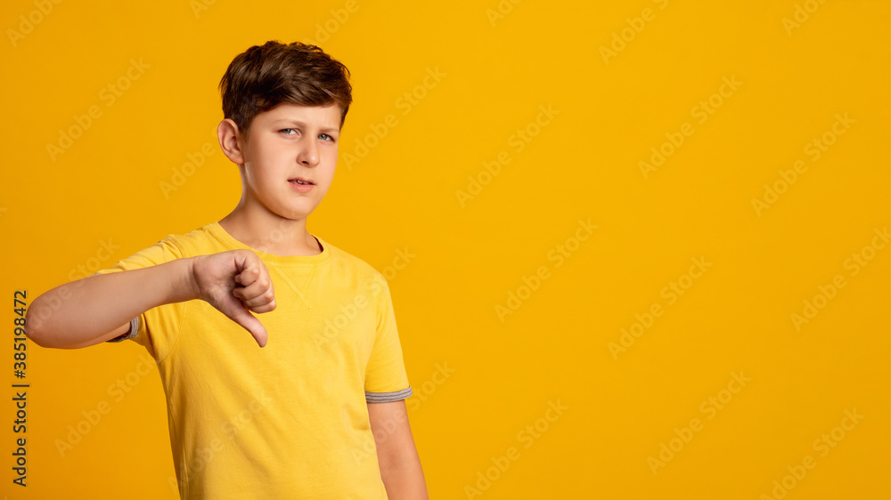 Dislike gesture. Disappointed kid. Wrong choice. Bad idea. Skeptic young boy rejecting offer with thumb down looking at camera isolated on orange copy space background. Dislike gesture. Disappointed kid. Wrong choice. Bad idea. Skeptic young boy rejecting offer with thumb down looking at camera isolated on orange copy space background.