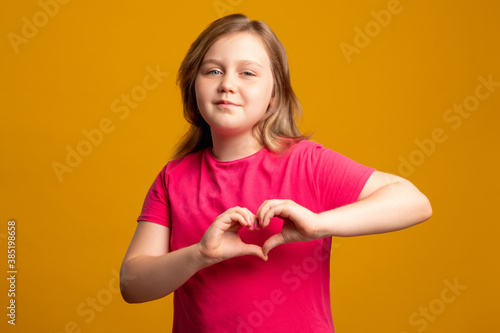 Love sign. Supportive kid. Admiration sympathy. Compassion encouragement. Confident young girl in pink showing heart gesture isolated on orange background.
