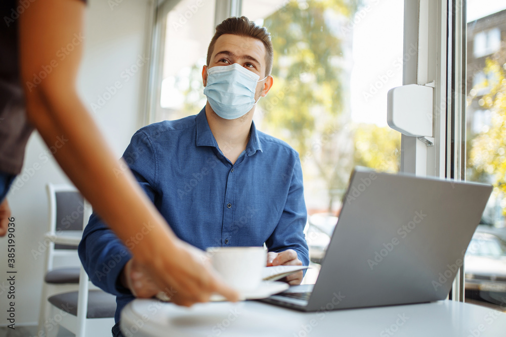 Waiter brings a cup of coffee to a young businessman wearing a sterile medical mask working from a cafe remotely. Distant business concept during coronavirus pandemic. Waiter brings a cup of coffee to a young businessman wearing a sterile medical mask working from a cafe remotely. Distant business concept during coronavirus pandemic.