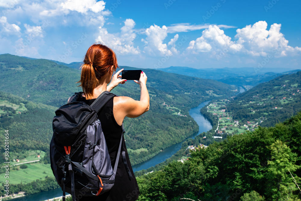 Rear view of female hiker with backpack standing on top of the mountain enjoying the view during the day and photographing landscape with phone.