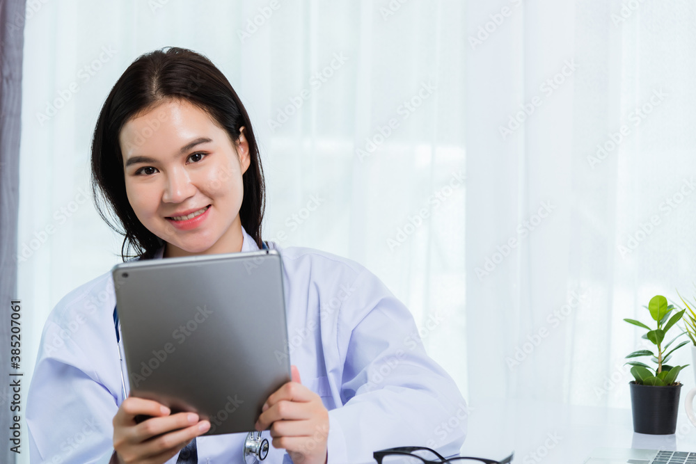 Asian young beautiful woman doctor on desk front laptop computer she holding modern smart digital tablet on hand during quarantines disease coronavirus or COVID-19 at hospital office Asian young beautiful woman doctor on desk front laptop computer she holding modern smart digital tablet on hand during quarantines disease coronavirus or COVID-19 at hospital office