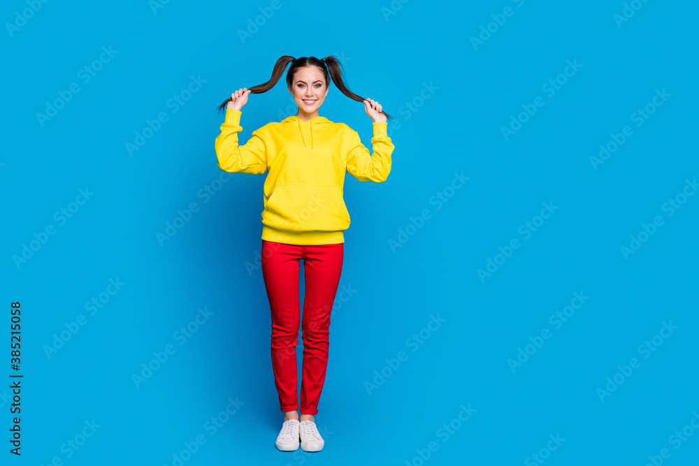 Full length photo of cheerful funny lady hold two tails arms good mood playful wear casual yellow hoodie pullover red trousers sneakers isolated bright blue color background Full length photo of cheerful funny lady hold two tails arms good mood playful wear casual yellow hoodie pullover red trousers sneakers isolated bright blue color background