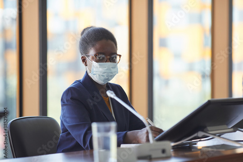 Portrait of African businesswoman in eyeglasses and in protective mask looking at camera while sitting at the table in board room
