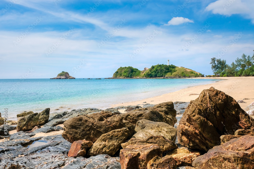 A beautiful tropical beach. Yellow sand and large stones in the foreground, turquoise sea and blue sky with clouds. A white lighthouse on a cliff in the distance. Koh Lanta, Thailand