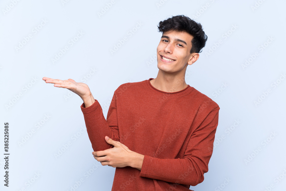 Young Argentinian man over isolated blue background extending hands to the side for inviting to come Young Argentinian man over isolated blue background extending hands to the side for inviting to come