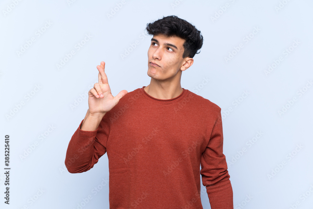 Young Argentinian man over isolated blue background with fingers crossing and wishing the best Young Argentinian man over isolated blue background with fingers crossing and wishing the best