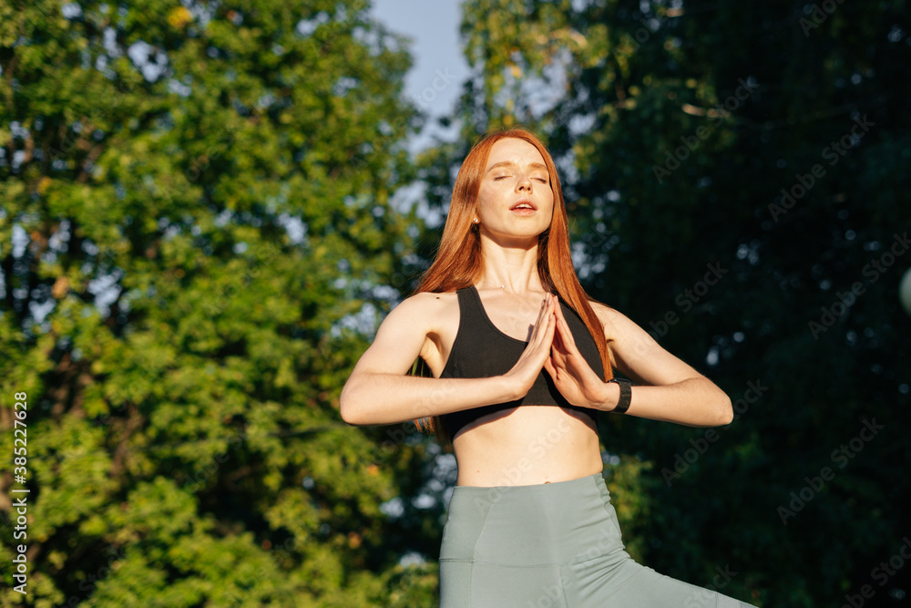 Low angle shot of attractive redhead young woman with closed eyes practicing yoga and meditation performing namaste pose. Calm female making Namaste gesture outside in city park in evening at sunset. Low angle shot of attractive redhead young woman with closed eyes practicing yoga and meditation performing namaste pose. Calm female making Namaste gesture outside in city park in evening at sunset.
