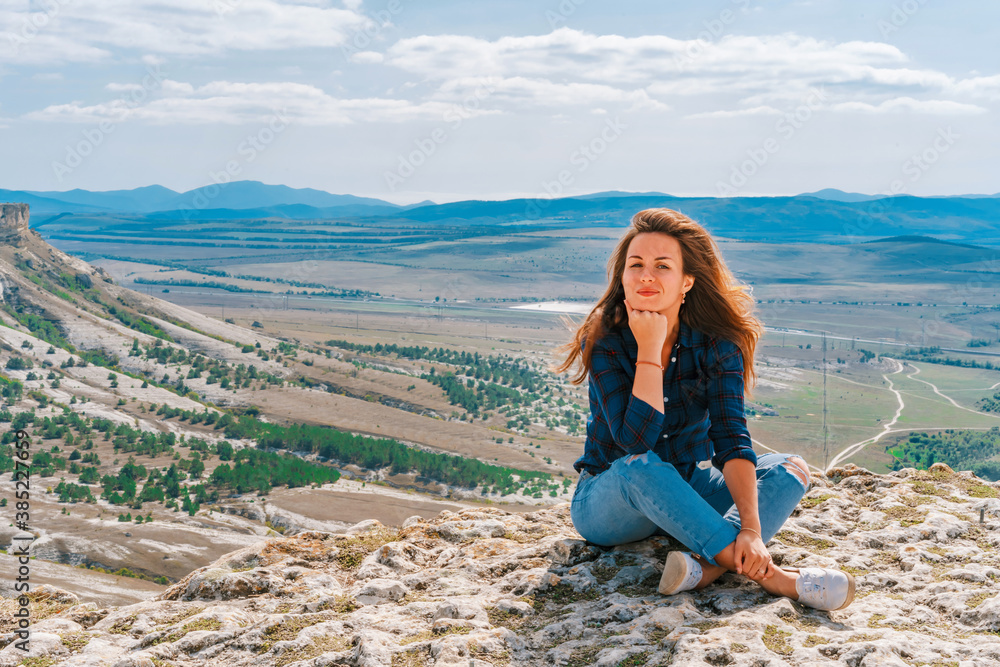 Beautiful young brunette woman in ripped jeans sitting on a cliff overlooking the rustic landscape, White rock in Crimea