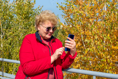 Caucasian senior woman in red jacket using smartphone, read e-book, leasten music or take online education in park in autumn weather Video call, chat Distance meeting  photo