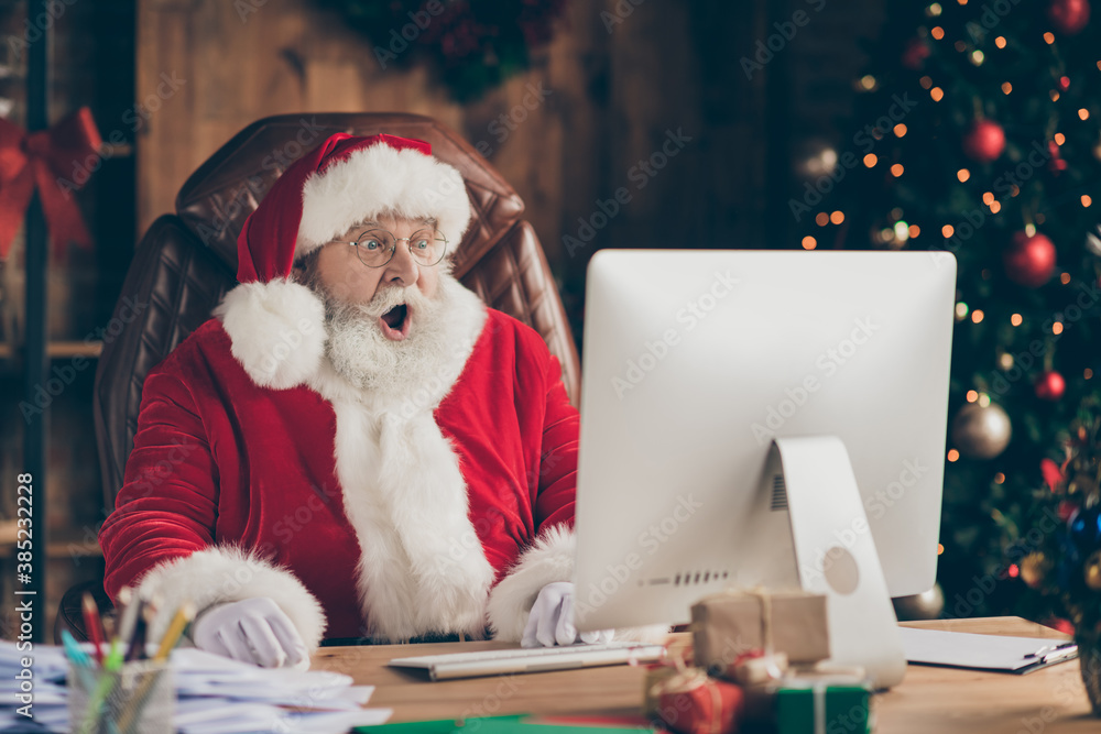 Photo of amazed santa claus sit table desk work computer impressed many  wish gift list letters in house idnoors with evergreen christmas x-mas tree  ornament decoration wear cap headwear Stock Photo