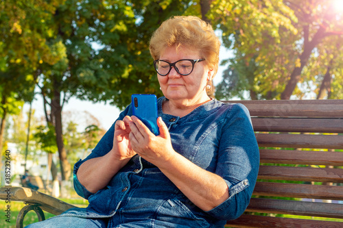 Senior woman using smartphone, read e-book, leasten music or take online education in park on bench in autumn weather Video call, chat Distance meeting  photo