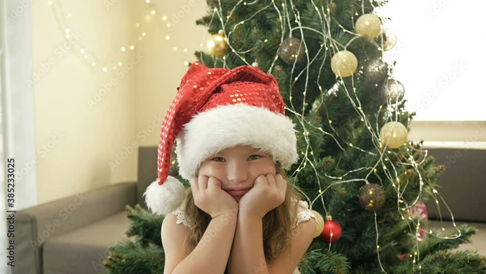 Portrait of a sad little girl in a Santa hat against the background of a Christmas tree. Portrait of a sad little girl in a Santa hat against the background of a Christmas tree.