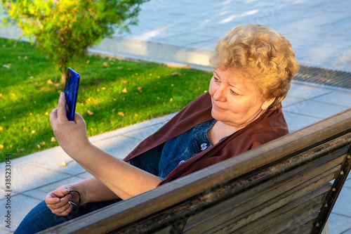 Senior woman using smartphone, read e-book, leasten music or take online education in park on bench in autumn weather in autumn weather Video call, chat Distance meeting 