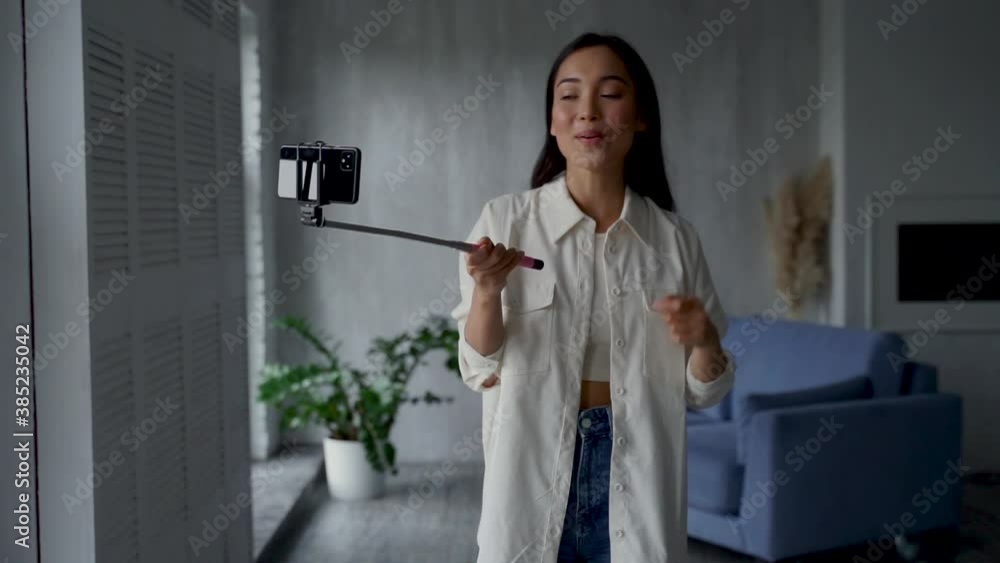 Female influencer broadcasting in streaming, doing online video call, she says goodbye waving hand and stops recording. Pretty asian young woman with a phone and a selfie stick, showing her home. Female influencer broadcasting in streaming, doing online video call, she says goodbye waving hand and stops recording. Pretty asian young woman with a phone and a selfie stick, showing her home.