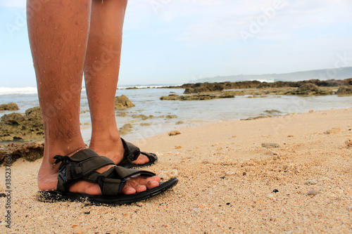 A pair of feet wearing black footwear is standing on the beach sand.
