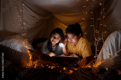 Family concept. elder sister and sister reading book with flashlight together before bedtime. Sister read story book together in bed sheet tent. focus selective little sister. With film grain effect © AKGK Studio