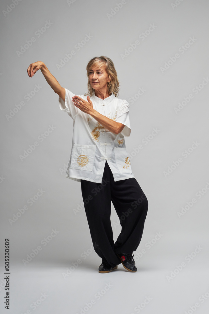 Mature blonde woman practicing Chi kung and Tai Chi on a white background. She wears a traditional white chinese Tai Chi jacket, black trousers and black shoes with ying yang symbol Mature blonde woman practicing Chi kung and Tai Chi on a white background. She wears a traditional white chinese Tai Chi jacket, black trousers and black shoes with ying yang symbol