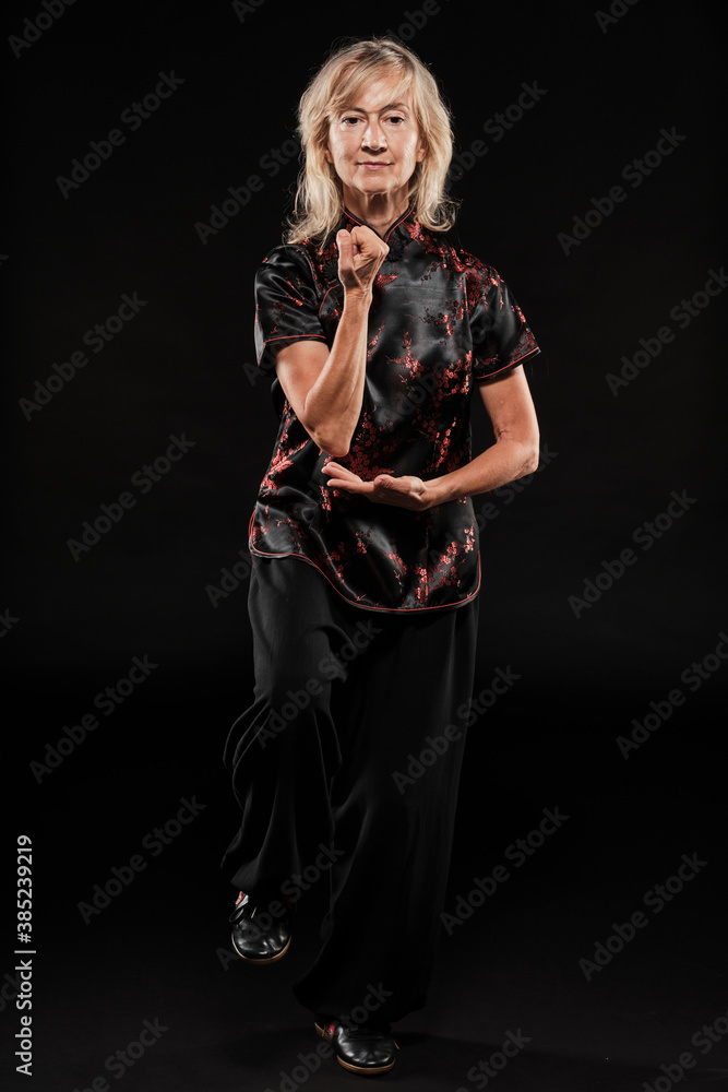 Mature blonde woman practicing Tai Chin Chen style in a black background wearing a traticional chinese black jacket with red decoration, black trousers and traditional shoes with ying yang symbol Mature blonde woman practicing Tai Chin Chen style in a black background wearing a traticional chinese black jacket with red decoration, black trousers and traditional shoes with ying yang symbol
