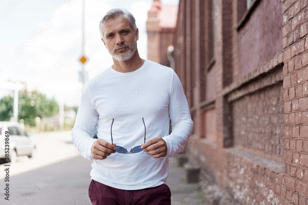 Obraz premium Portrait of a Grey-haired Mature handsome man in jeans and white t-shirt leaning to the old wall.