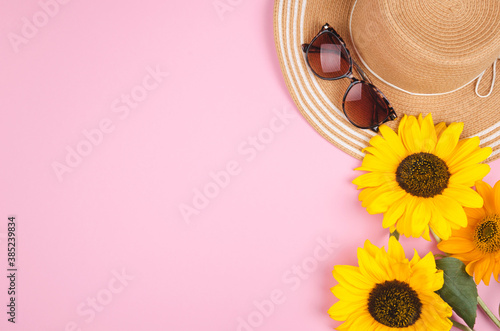 Flatlay with sunglasses, straw hat and bright yellow sunflower on pink background.