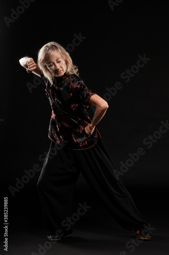 Mature blonde woman practicing Tai Chin Chen style in a black background wearing a traticional chinese black jacket with red decoration, black trousers and traditional shoes with ying yang symbol